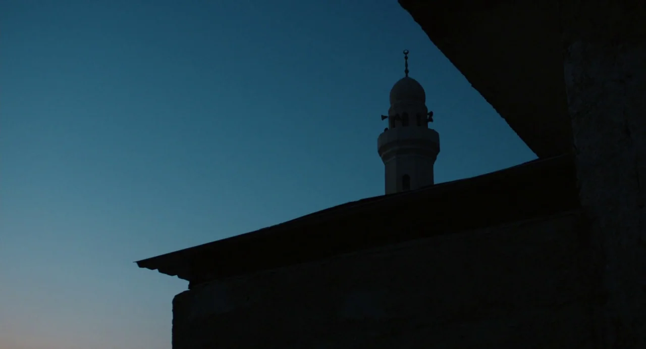 Zero Dark Thirty (2012) movie still: establishing — A minaret rises above a dark rooftop silhouetted against a deep blue dusk sky. The building…