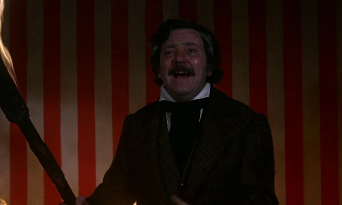 Vampire Circus (1972) movie still: medium — A man stands before a backdrop of vertical red and white stripes. He holds a wooden torch while speaking…