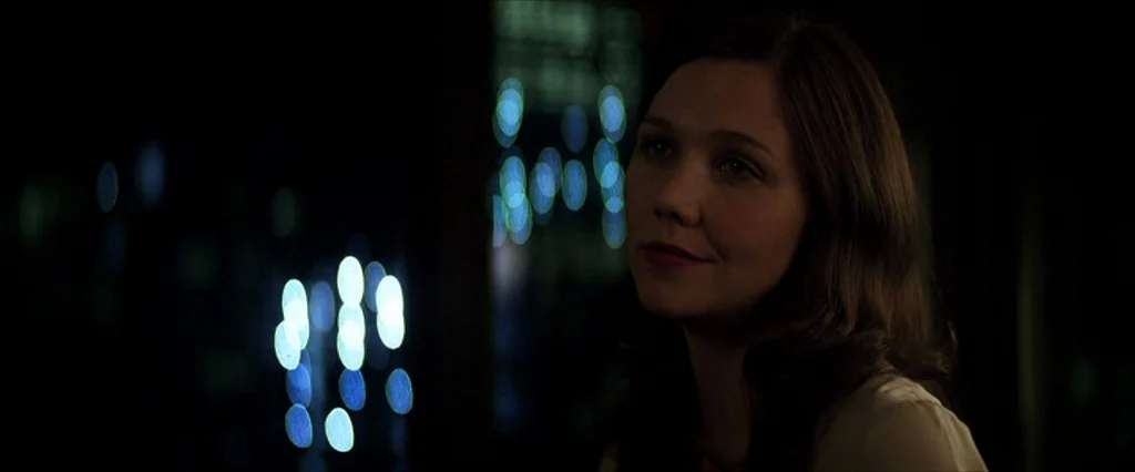 The Dark Knight (2008) movie still: medium — A dark indoor space is filled with blurry blue bokeh lights from an exterior view. A woman sits in the…