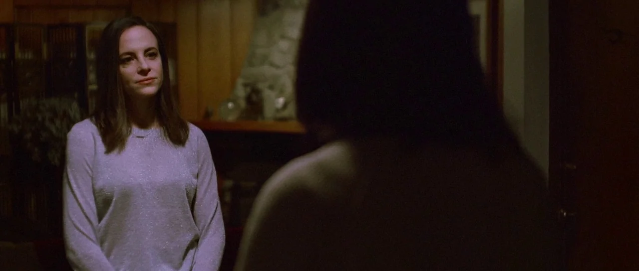 Depraved (2019) movie still: medium — A dimly lit room with wood-paneled walls and a stone fireplace in the back. A woman stands in the foreground…