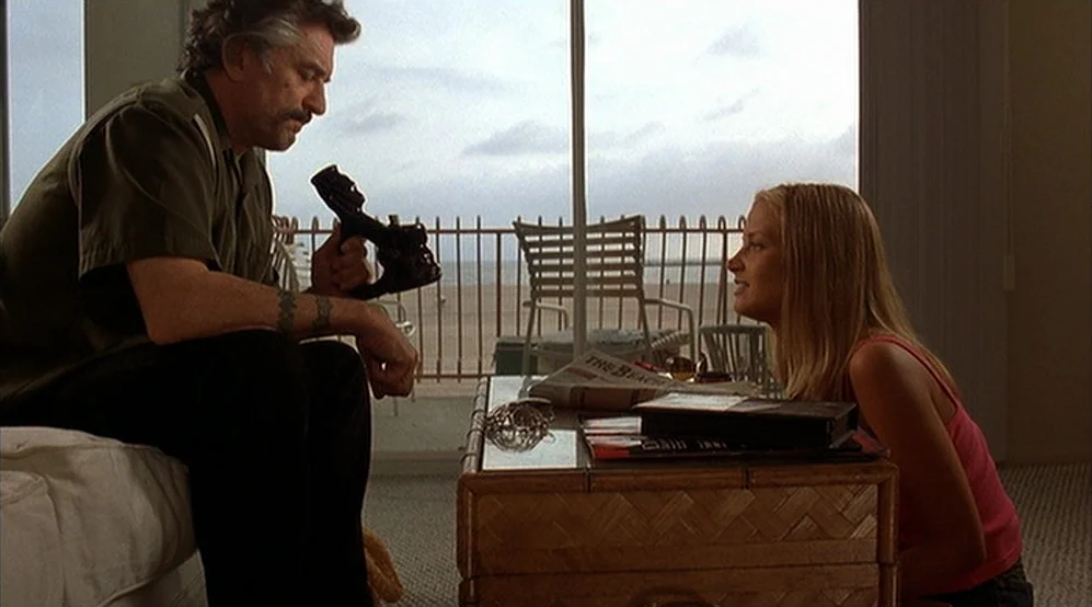 Jackie Brown movie still: medium — A hotel room with a glass door overlooking a beach. A man sits on a bed while a woman faces him across a table.