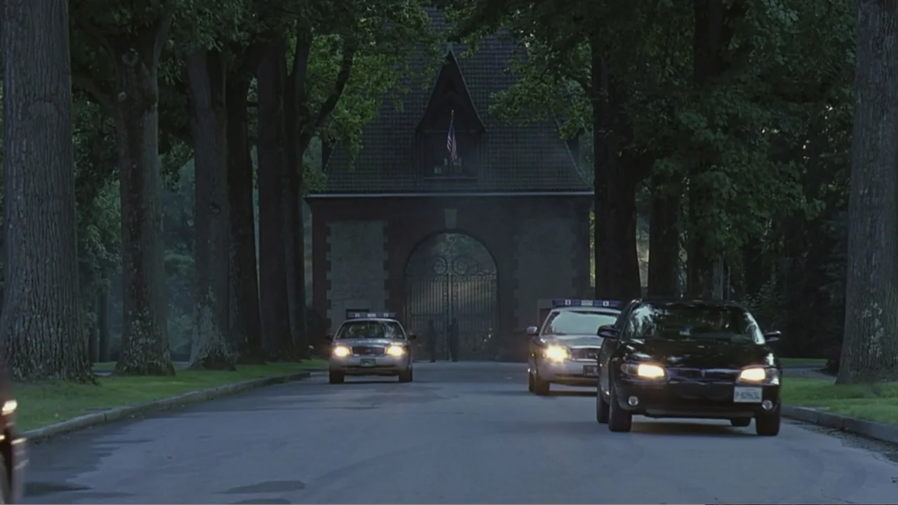 Hannibal (2001) movie still: wide — A paved driveway is lined with tall mature trees leading toward an ornate gate. Three police vehicles are…