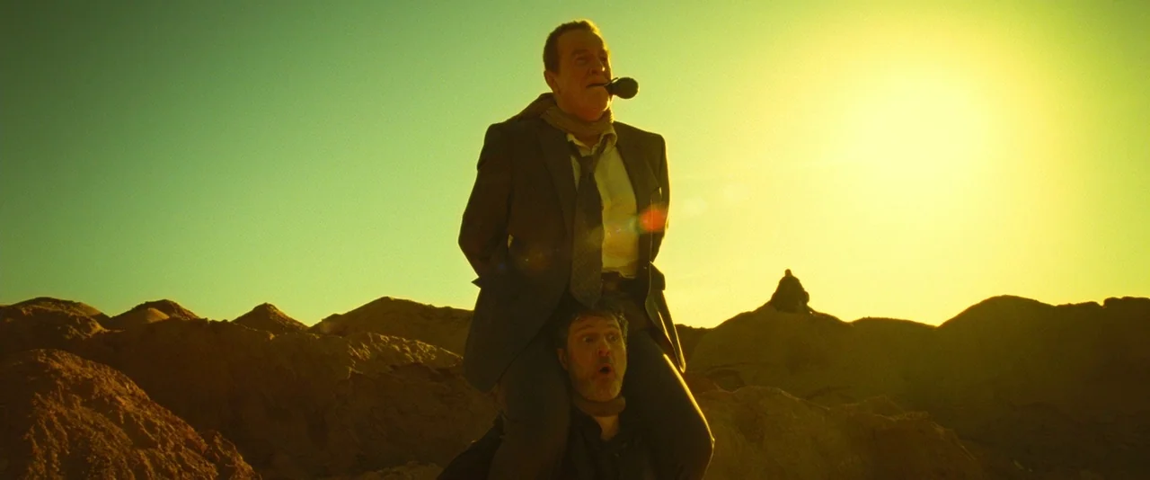 Micmacs (2009) movie still: fullbody — A vast, uneven, yellow-hued desert terrain stretches under a clear, bright sky. One man is sitting on the…