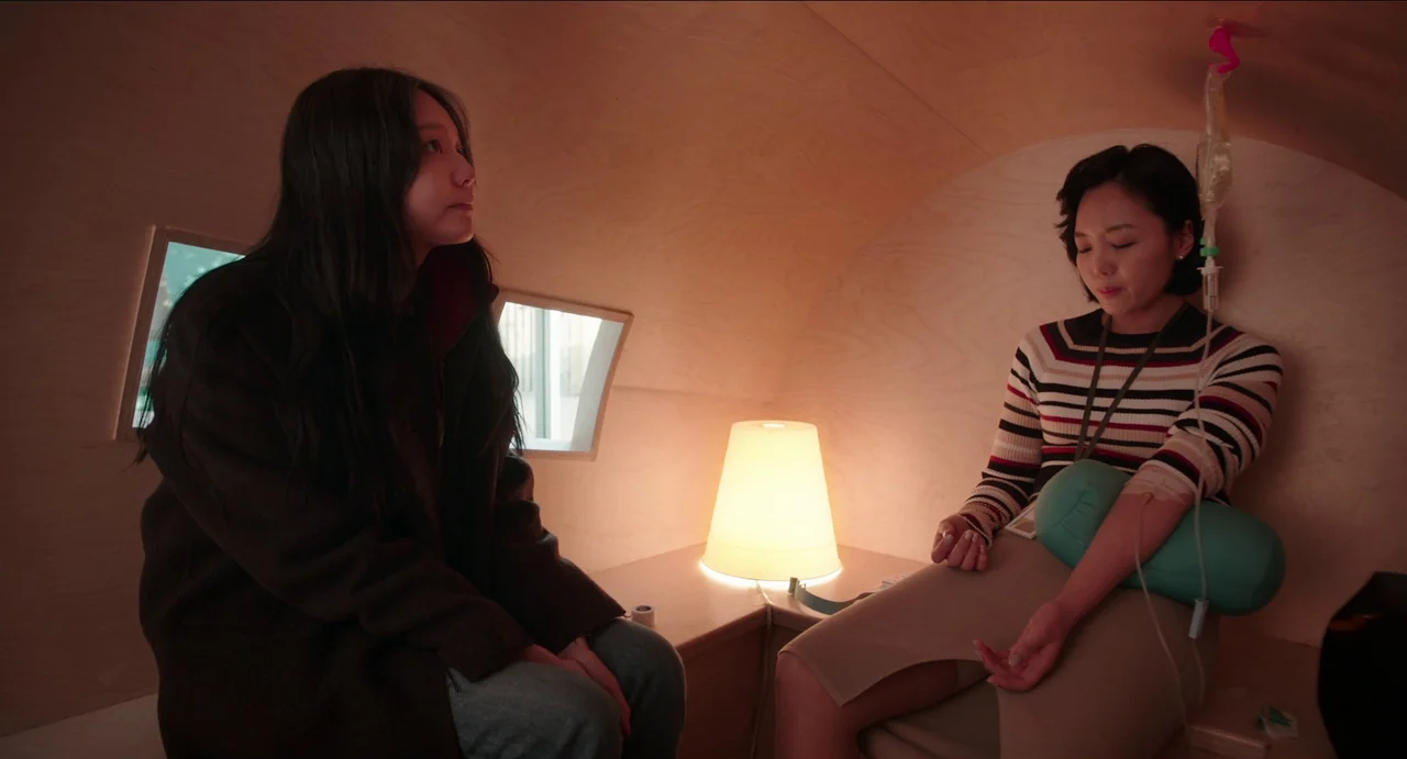 Microhabitat (2017) movie still: medium — A small room features curved plywood walls and two small square windows set deep. One woman sits watching…