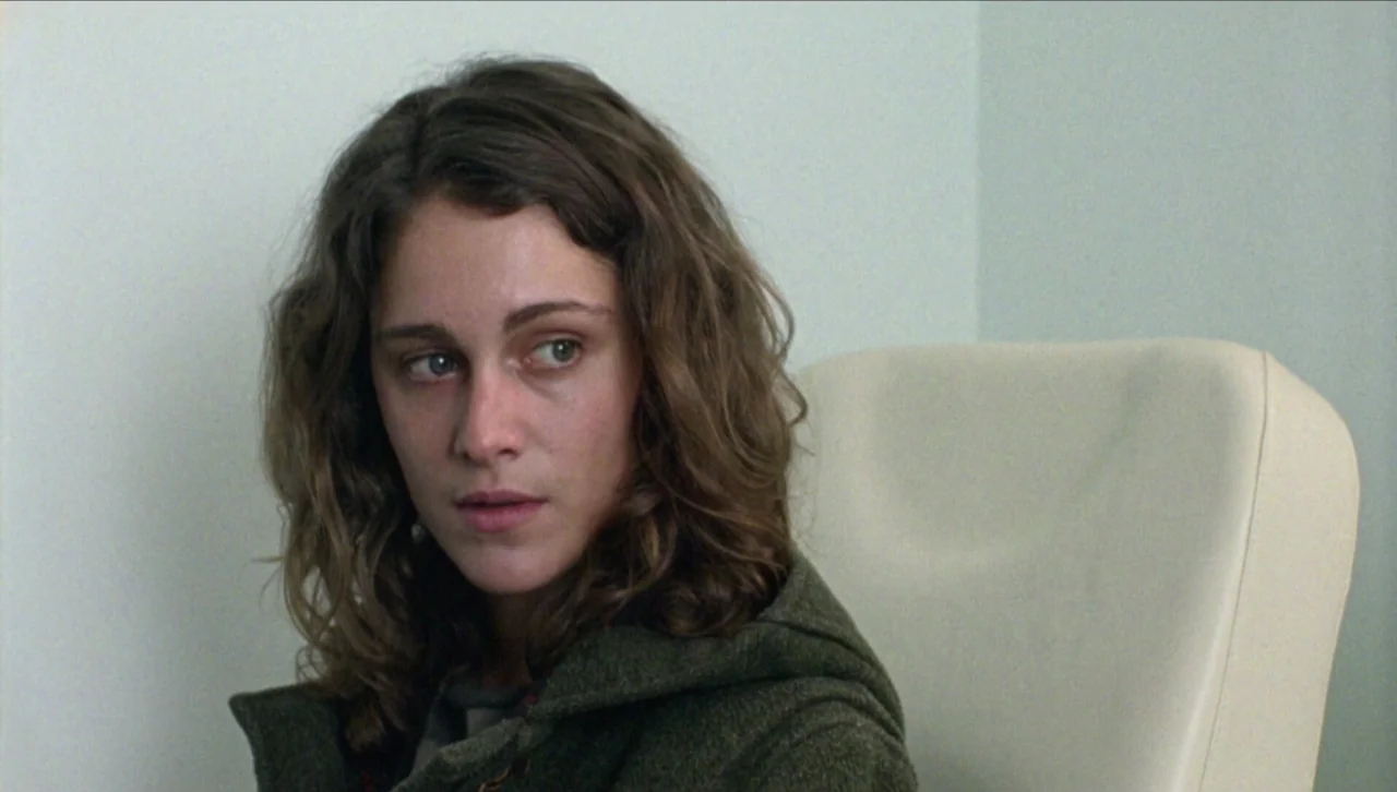 Attenberg (2010) movie still: closeup — A woman sits in a medical office with pale light-green walls. She is sitting in a white examination chair…
