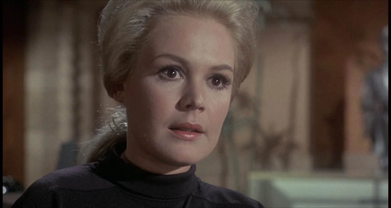 The Dunwich Horror (1970) movie still: closeup — A dimly lit indoor office setting with blurred architectural details in the background. A woman…