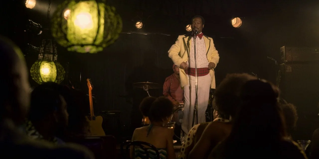Dolemite Is My Name movie still: medium — A dimly lit club features patrons in the foreground facing a small stage area. The performer stands…