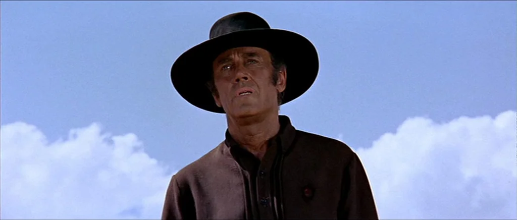 Once Upon A Time In The West (1968) movie still: medium — A man stands outdoors under a bright blue sky filled with scattered white clouds. The man…