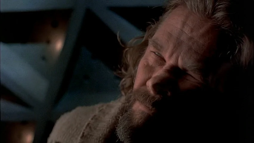 The Big Lebowski (1998) movie still: closeup — A man with a beard sits in a darkened, indistinct interior space. His face is illuminated by a…