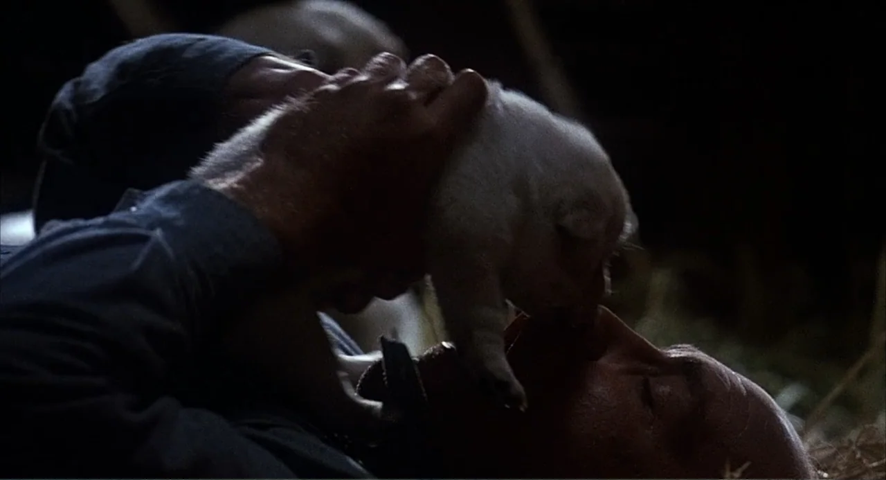 Of Mice and Men (1992) movie still: closeup — A dark interior space with straw visible in the low-light background. A man lies down while lifting a…