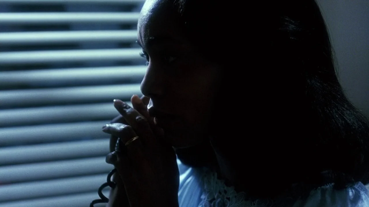 Malcolm X (1992) movie still: closeup — A woman stands in a dimly lit room against closed horizontal blinds at night. She holds a black telephone…