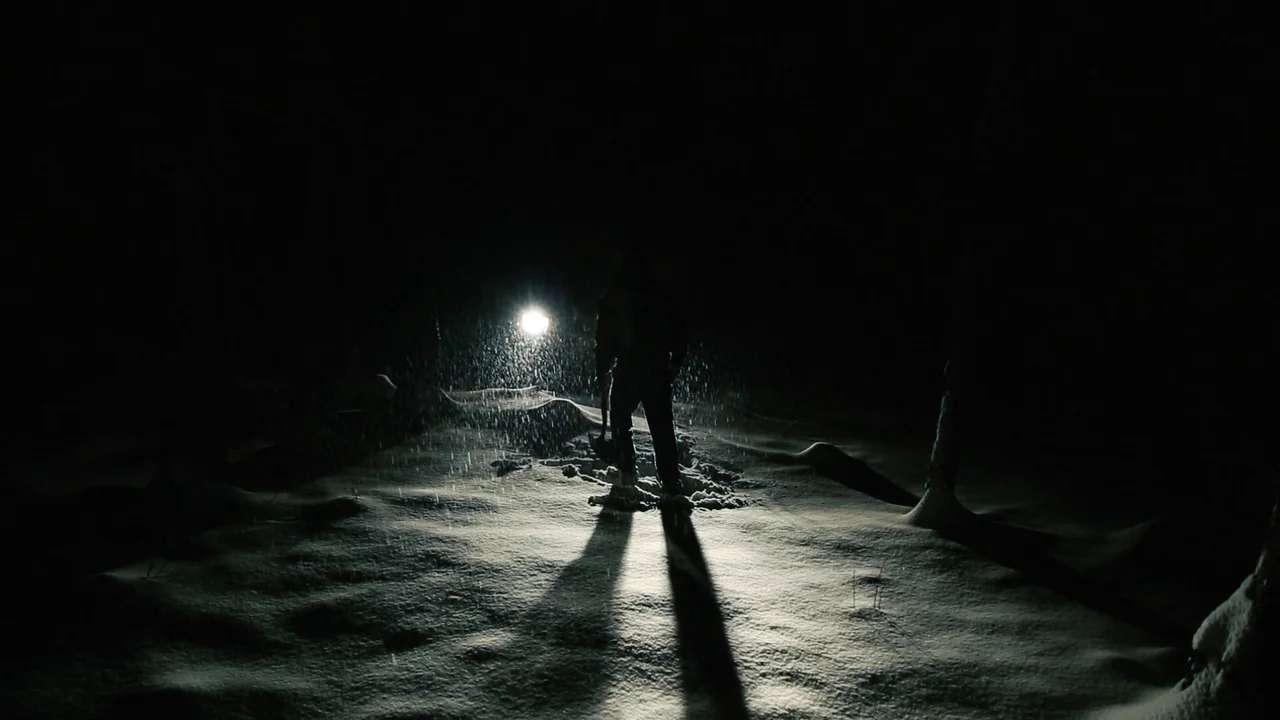 The Deeper You Dig (2019) movie still: fullbody — A dark snowy landscape at night is illuminated by a single bright light source. A solitary person…