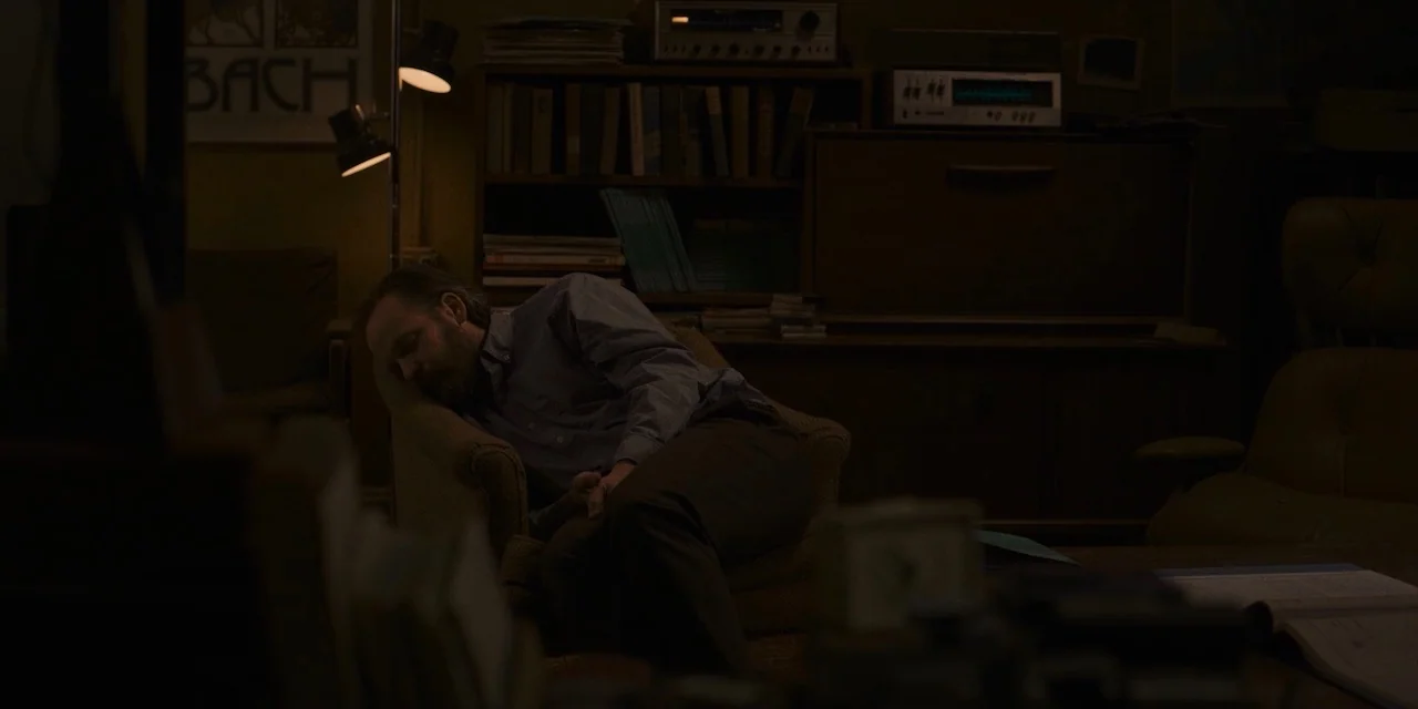 The Sound of Silence movie still: medium — A dimly lit room contains wooden shelves filled with books and vintage audio equipment. A man sleeps in a…