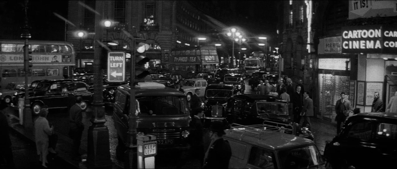 Bunny Lake Is Missing (1965) movie still: wide — A crowded London street intersection at night filled with vintage cars and double-decker buses.…