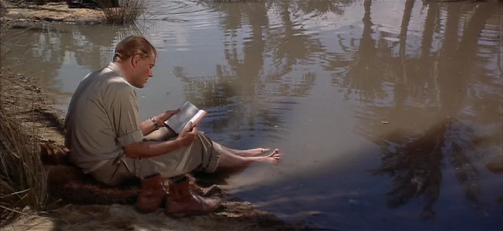 Lawrence Of Arabia movie still: wide — A remote oasis in a desert landscape features a small pool reflecting palm trees. A man in tan clothing sits…