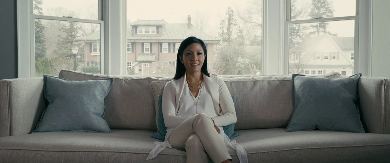 Hustlers (2019) movie still: medium — A woman sits on a beige sofa positioned in front of a bay window. Through the glass, a two-story residential…