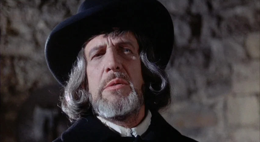 Witchfinder General (1968) movie still: closeup — A man stands before a rough-hewn stone wall in a dimly lit enclosure. He stares forward with an…