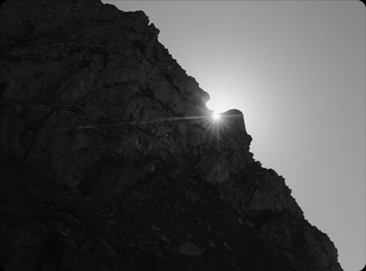 Eureka (2024) movie still: wide — A dark rocky mountain ridge silhouettes against a bright clear sky. The sun bursts over the jagged edge of the…
