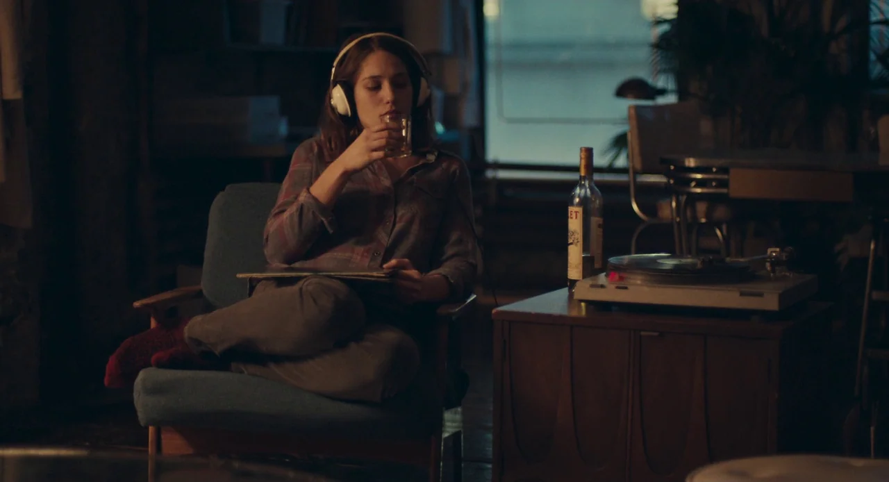 Mistress America (2015) movie still: medium — A dimly lit room contains a blue armchair and a wooden record player stand. A woman sits in the chair…
