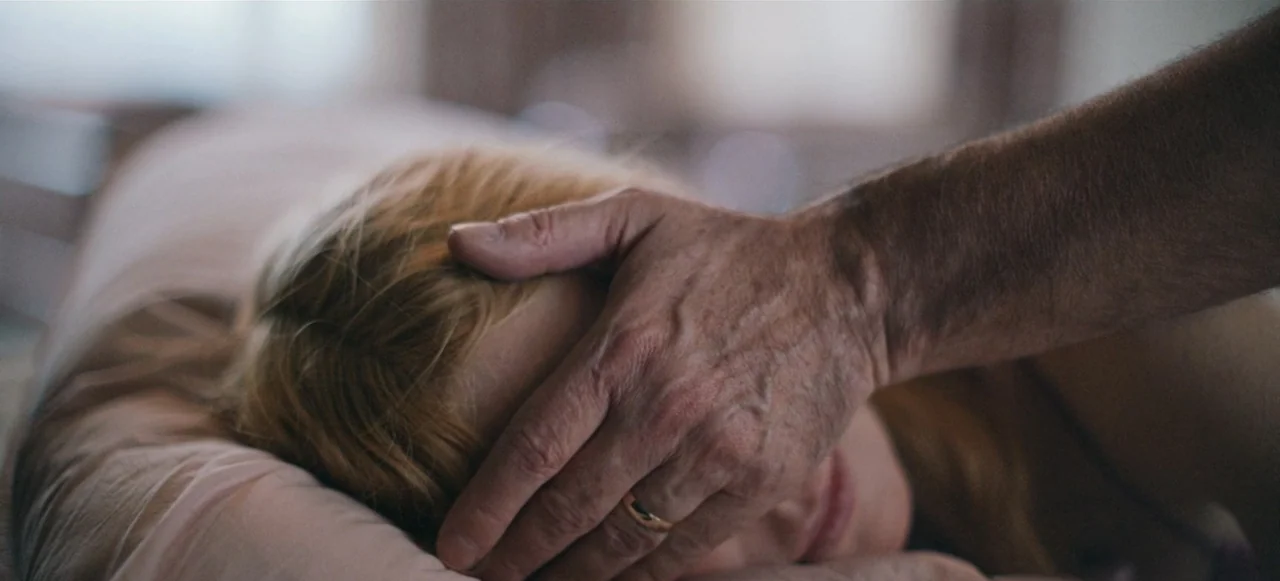 Babygirl (2024) movie still: closeup — A dimly lit bedroom shows a person resting on a pillow. A man's hand reaches out to touch the forehead of the…