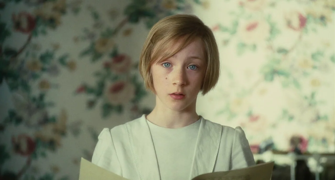 Atonement (2007) movie still: medium — A room with vintage floral wallpaper features a young girl centered in the frame. The girl looks forward…