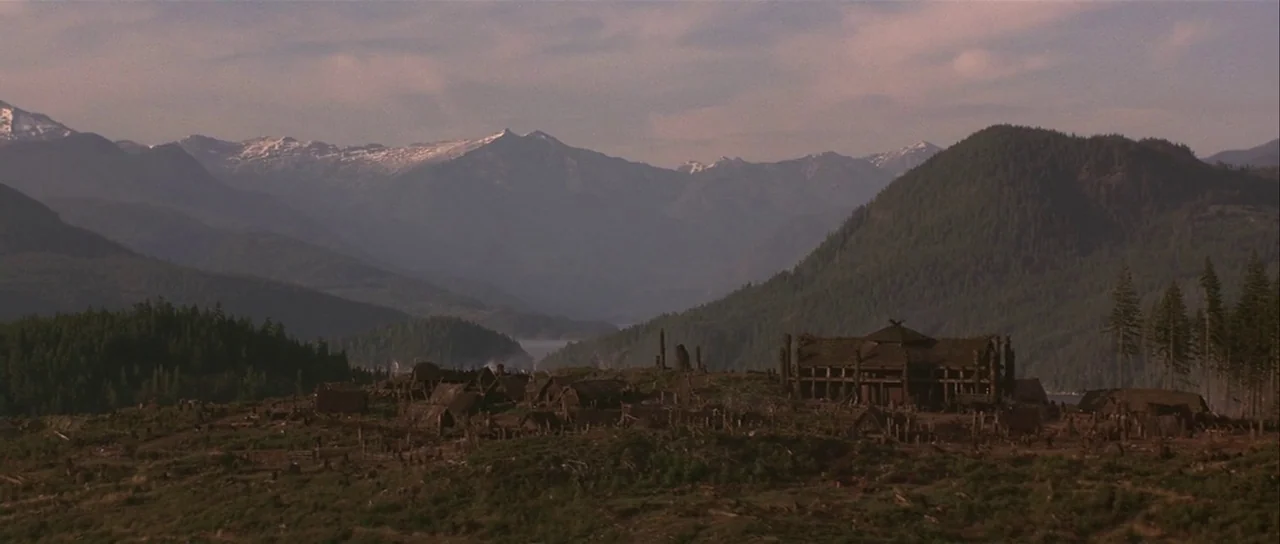 The 13th Warrior (1999) movie still: establishing — A collection of wooden longhouses sits on a clearing surrounded by dense coniferous forests.…