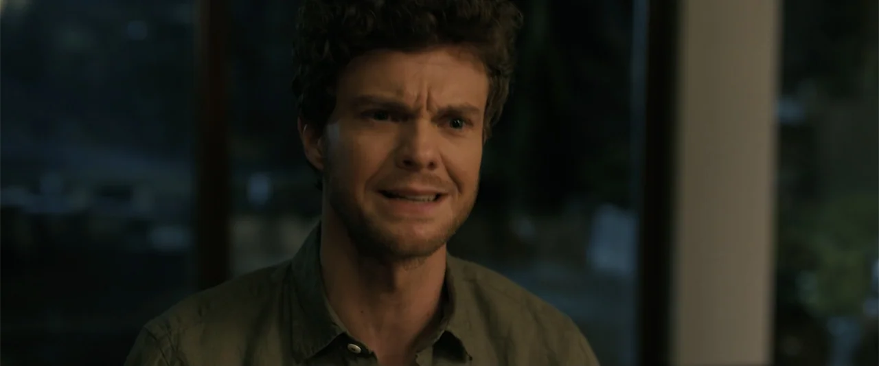 Companion (2025) movie still: medium — A dimly lit indoor room serves as the backdrop for a close subject. A young man with brown curly hair looks…