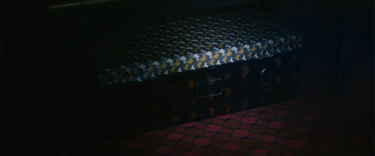 Ms. Kanyin (2025) movie still: closeup — A dark, dimly lit corner of an interior space features a vintage rectangular trunk. The trunk sits on a red…