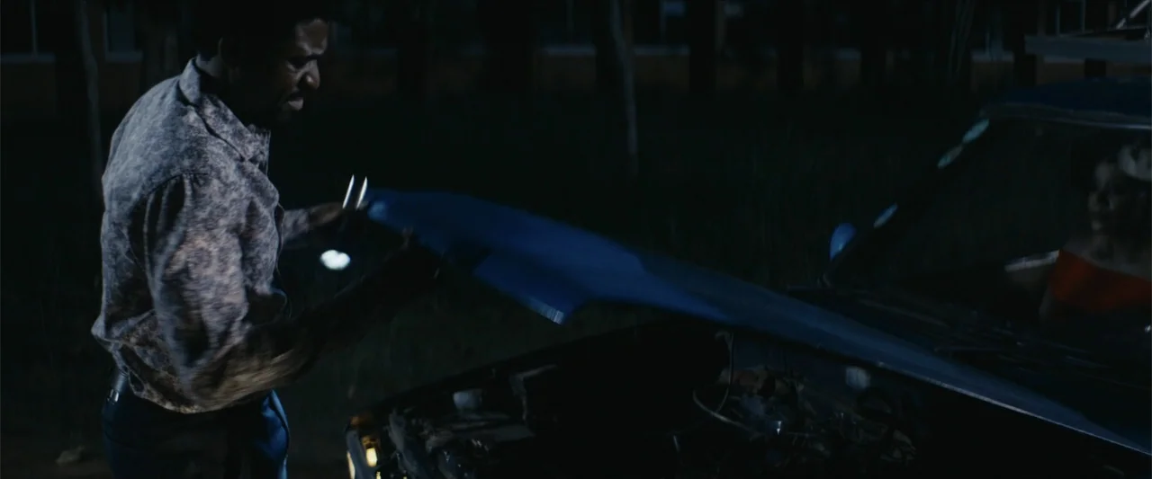 Ms. Kanyin (2025) movie still: medium — A dark roadside at night features a man inspecting an open car hood. The vehicle is parked outdoors with…