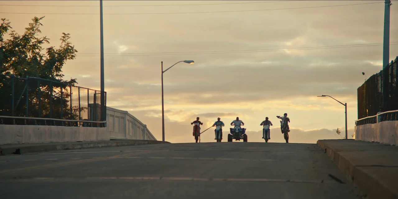 Charm City Kings (2020) movie still: wide — A paved bridge spans the frame under a cloudy sunset sky with silhouette lighting. Five riders on…