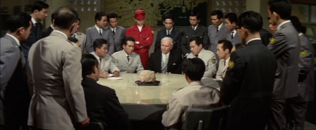 Destroy All Monsters (1968) movie still: wide — A group of men in military uniforms and suits surround a circular table inside a room. An…