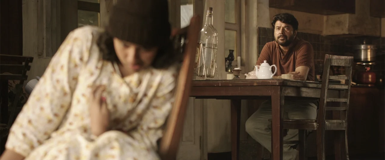 Peranbu (2019) movie still: medium — A domestic space with a wooden dining table and blurred background features. A man in a brown shirt watches a…