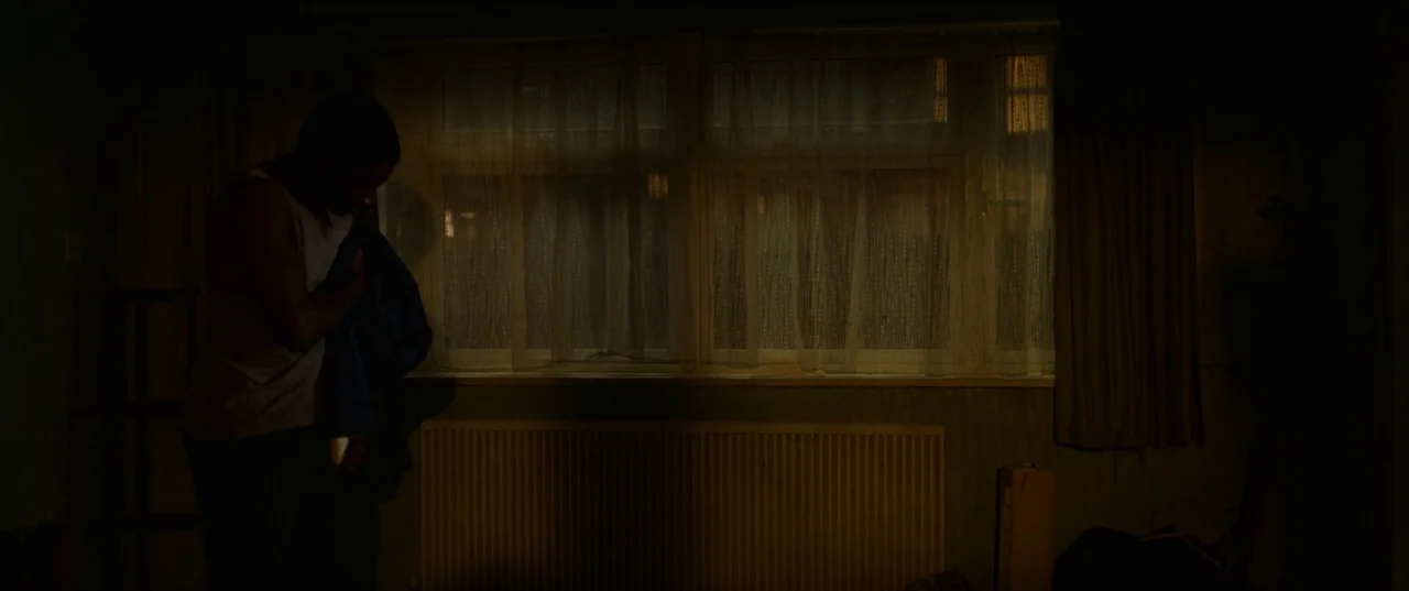 His House (2020) movie still: medium — A dark, dimly lit room features a window covered by thin, semi-transparent curtains. A man stands in profile…