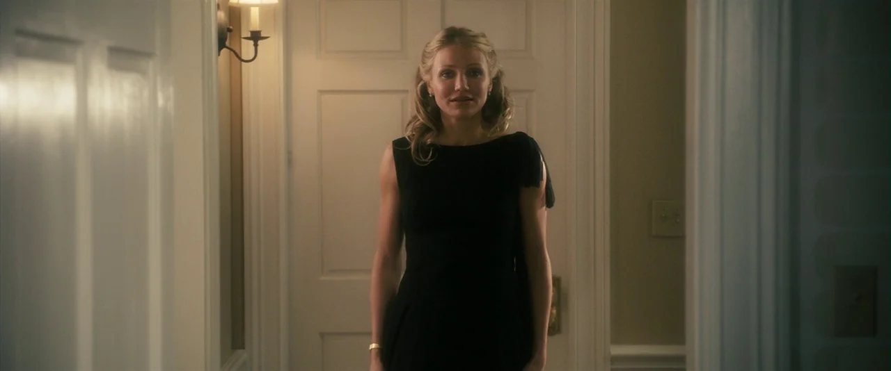 The Box (2009) movie still: medium — A residential hallway with white panelled doors and off-white walls. A woman in a black dress stands in the…
