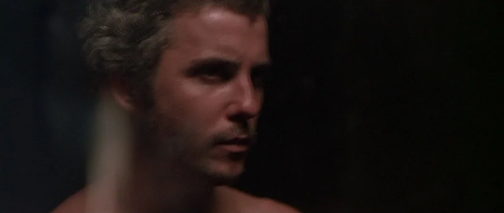Manhunter (1986) movie still: closeup — A man stands in a dark, indistinct interior setting with minimal environmental detail. The man is positioned…