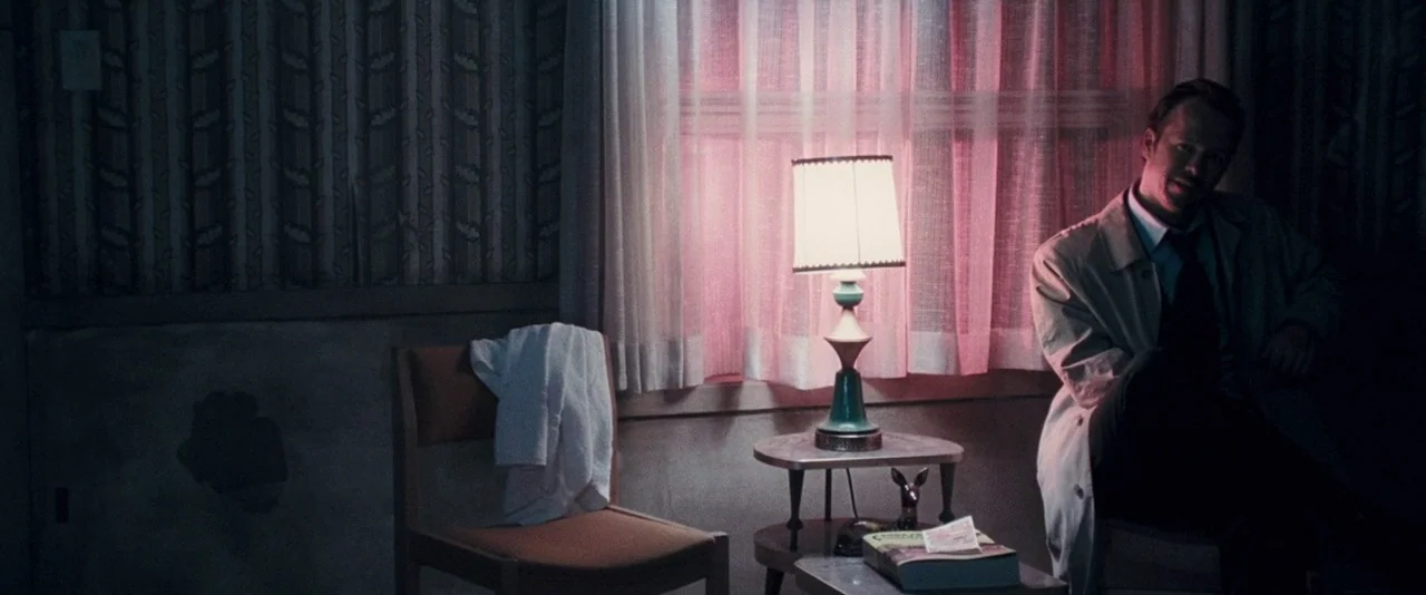 Dead Silence (2007) movie still: medium — A dark, dimly lit motel room features patterned wallpaper and a small lamp. A man wearing a trench coat…