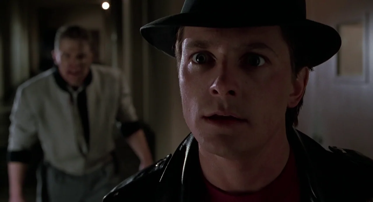 Back to the Future II (1989) movie still: closeup — A dark, narrow interior hallway with blurred walls in the background. A man in a leather jacket…