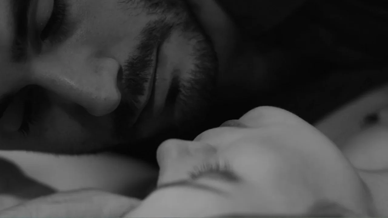 Creditors (2025) movie still: closeup — A dark bedroom interior features two people lying closely on a bed. The man's face is positioned near the…
