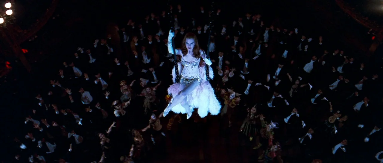Moulin Rouge! (2001) movie still: wide — A crowded, dimly lit 19th-century performance hall with balconies and dark wood architecture. A woman sits…