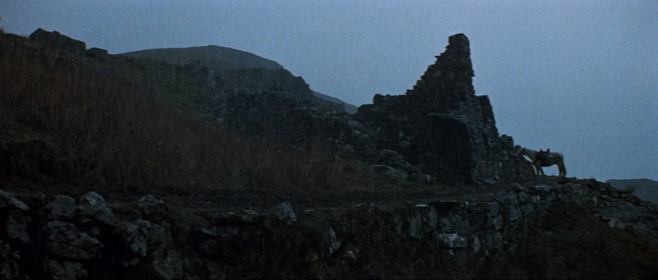 Macbeth (1971) movie still: wide — An isolated set of stone ruins sits on a grassy, rocky hillside under dusk. A lone horse stands saddled beside…