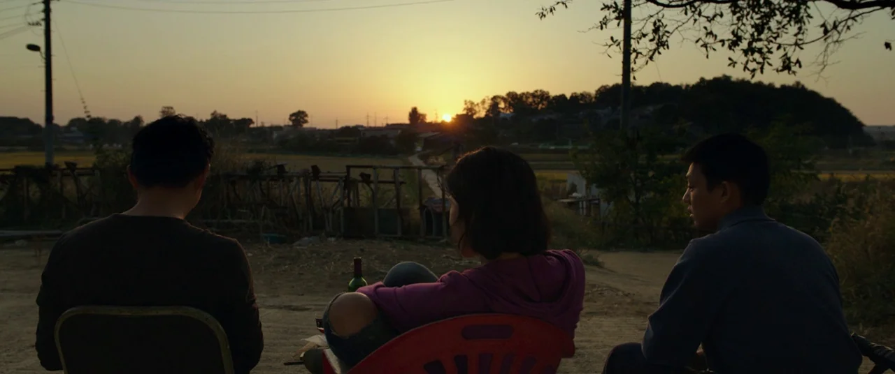 Burning (2018) movie still: wide — Three people sit on plastic chairs on a dusty road facing a sunset. Utility poles and vegetation border the open…