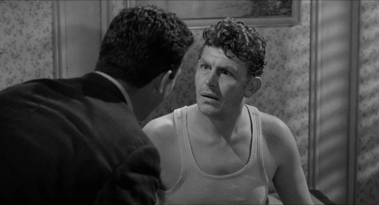 A Face In The Crowd (1957) movie still: medium — A man wearing a tank top sits in a bedroom with patterned wallpaper. He stares intently at a man in…