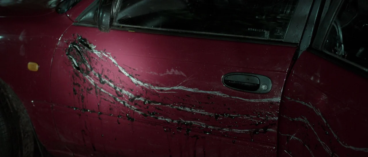 The Hallow (2015) movie still: closeup — A maroon car door features severe, deep gashes running across the metal surface. Thick black fluid drips…