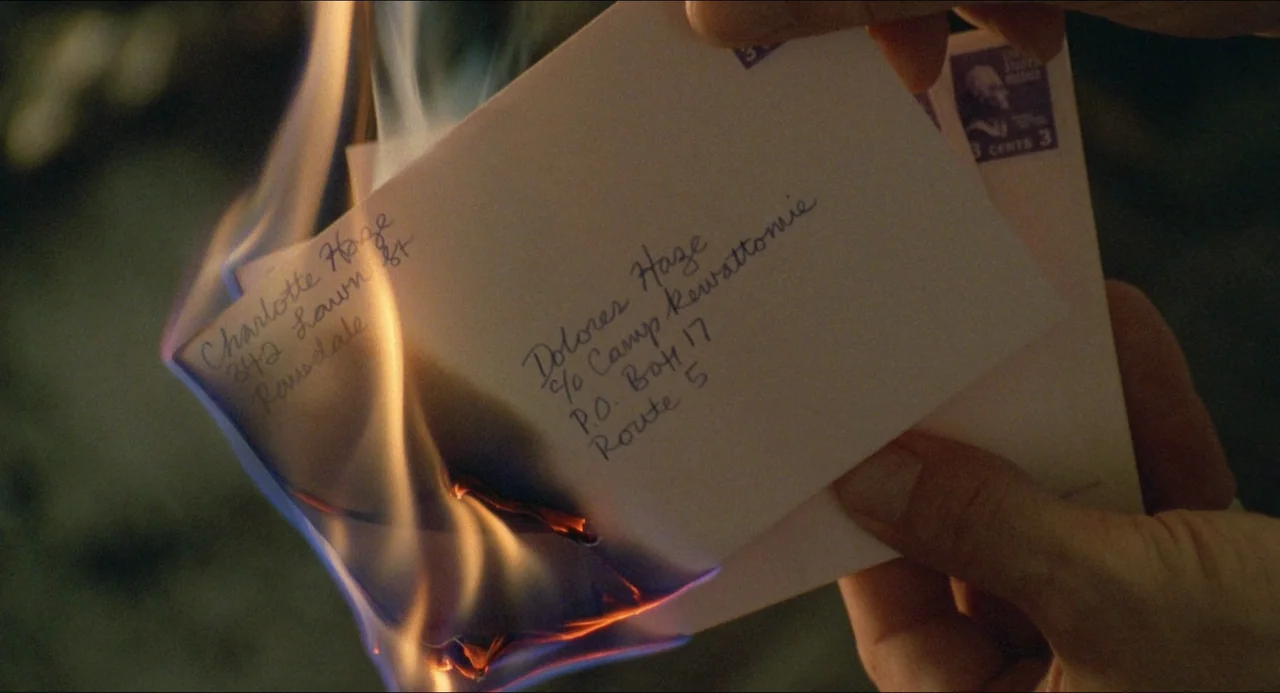Lolita (1997) movie still: closeup — A close-up shows a white paper envelope being consumed by active orange flames. Cursive ink handwriting…