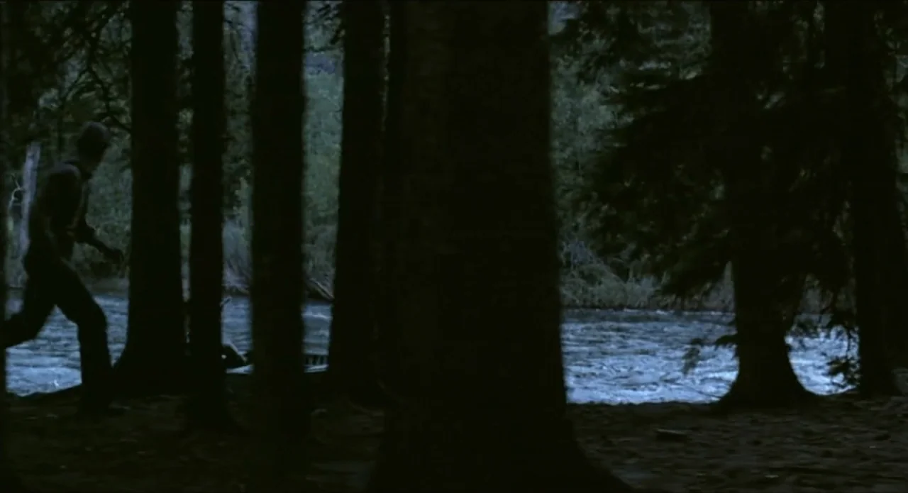 A River Runs Through It (1992) movie still: medium — A dark riverbank is framed by dense pine trees at night. A lone man walks along the edge of the…