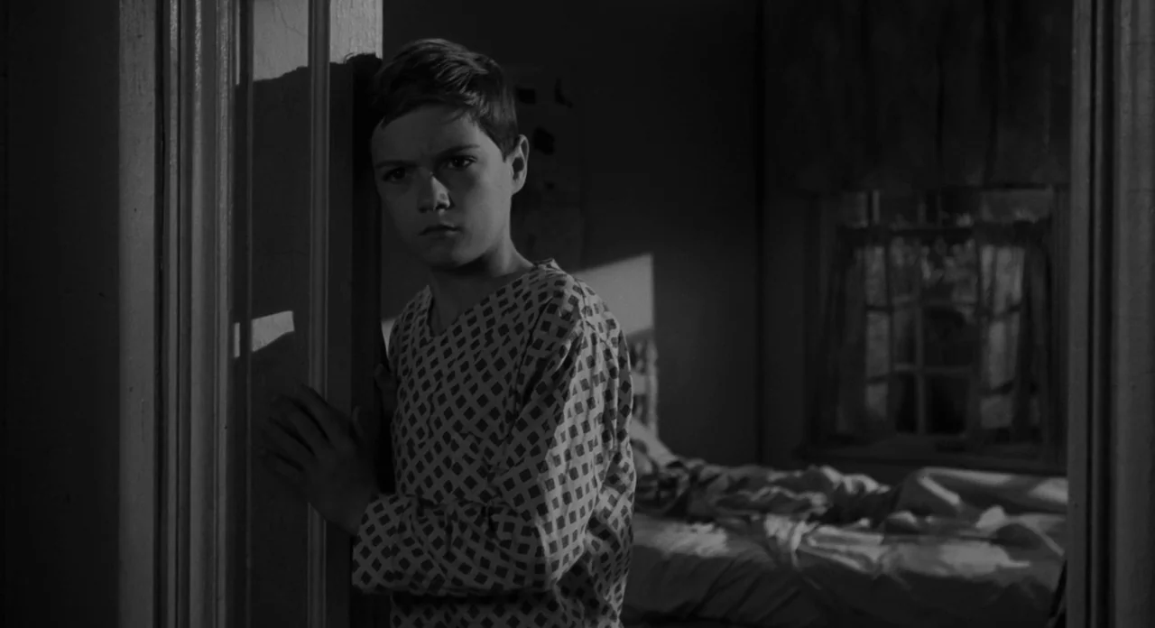 To Kill A Mockingbird (1962) movie still: medium — A dark, sparsely lit room with a window visible in the background. A young boy in pajamas stands…