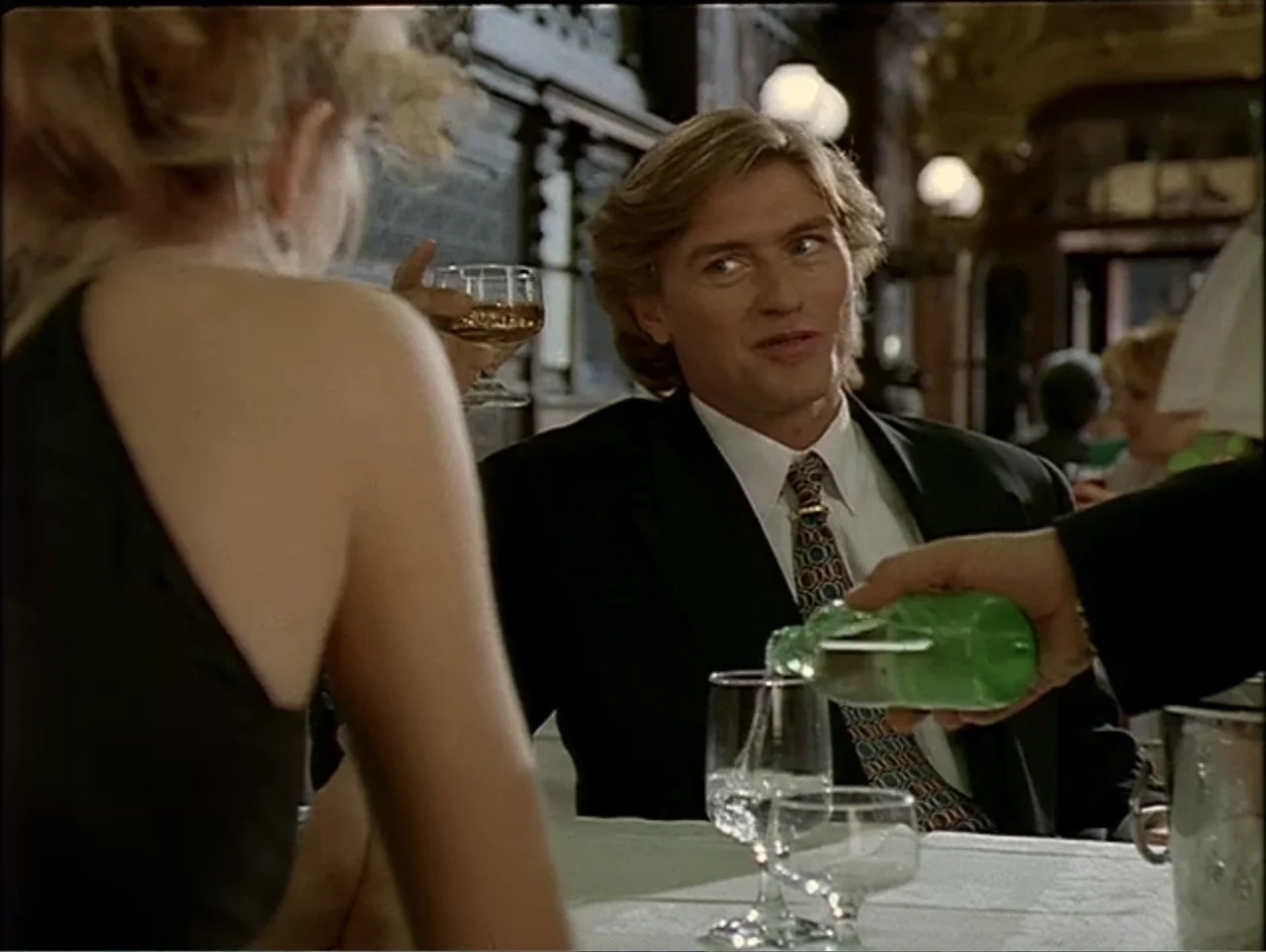 A Change of Place (1994) movie still: medium — A restaurant interior features white tablecloths and dark paneled walls in the background. A man sits…