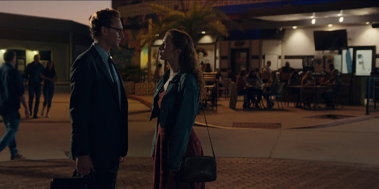 The Life of Chuck (2024) movie still: medium — An outdoor paved area during twilight features people dining at tables in the background. A man in a…