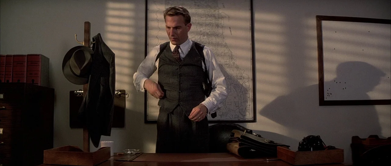 The Untouchables (1987) movie still: medium — A 1930s-style room features a wooden desk, a wall map, and a hat rack. A man in a suit adjusts his…