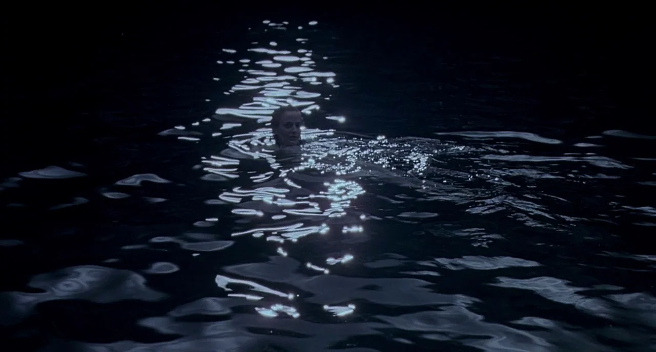 Cracks (2009) movie still: wide — Dark, calm water stretches across the frame under a night sky. A person's head and shoulders are visible above the…